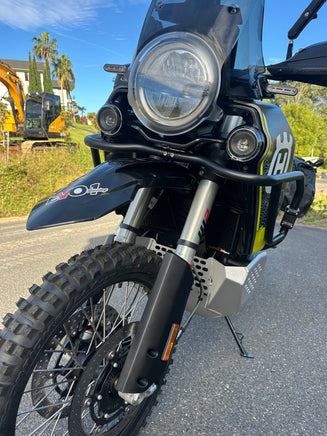 Close-up front view of a Husqvarna off-road motorcycle parked on an asphalt road, featuring a black front fender with spacer kit installed from RAD GUARD, round headlight, black front bumper guard, and knobby tires.